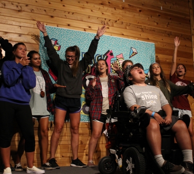 A group of more than 10 activity leaders sing and dance in the background for a camper singing at a MDA Camp's talent show.