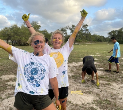 Gillian and Anna throw arms out wide and smile at the camera with paint covered faces. campers are in the background picking up marshmallows form the paint war