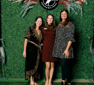 Lillian, Kate, and Mandy stand in front of a greenery wall that has a camp aranzazu logo in the center above their heads
