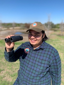 A Camp Aranzazu activity leader, Angel, smiles holding binoculars while wearing the olive camp hat and embroidered flannel.