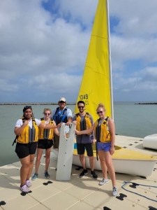 two activity leaders stand on each side of Ted as they pose with a piece of steering equipment and in front of the sailboat on copano bay. ted and activity leaders are wearing life jackets and smiling at the camera
