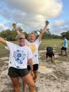 Gillian and Anna throw arms out wide and smile at the camera with paint covered faces. campers are in the background picking up marshmallows form the paint war