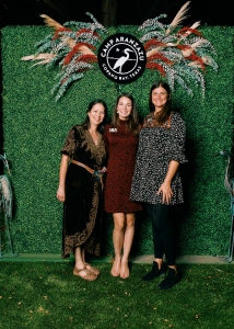 Lillian, Kate, and Mandy stand in front of a greenery wall that has a camp aranzazu logo in the center above their heads